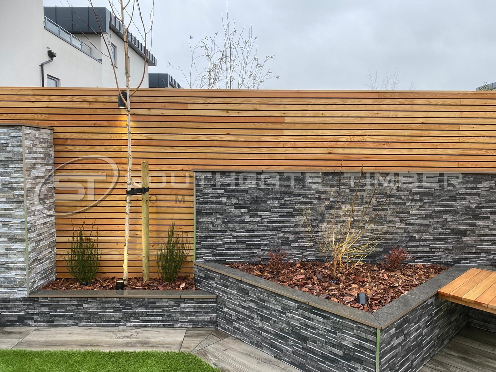 Contemporary garden design featuring European Larch horizontal slatted fencing with warm golden tones, paired with dark stone raised planters and seating, supplied by Southgate Timber.