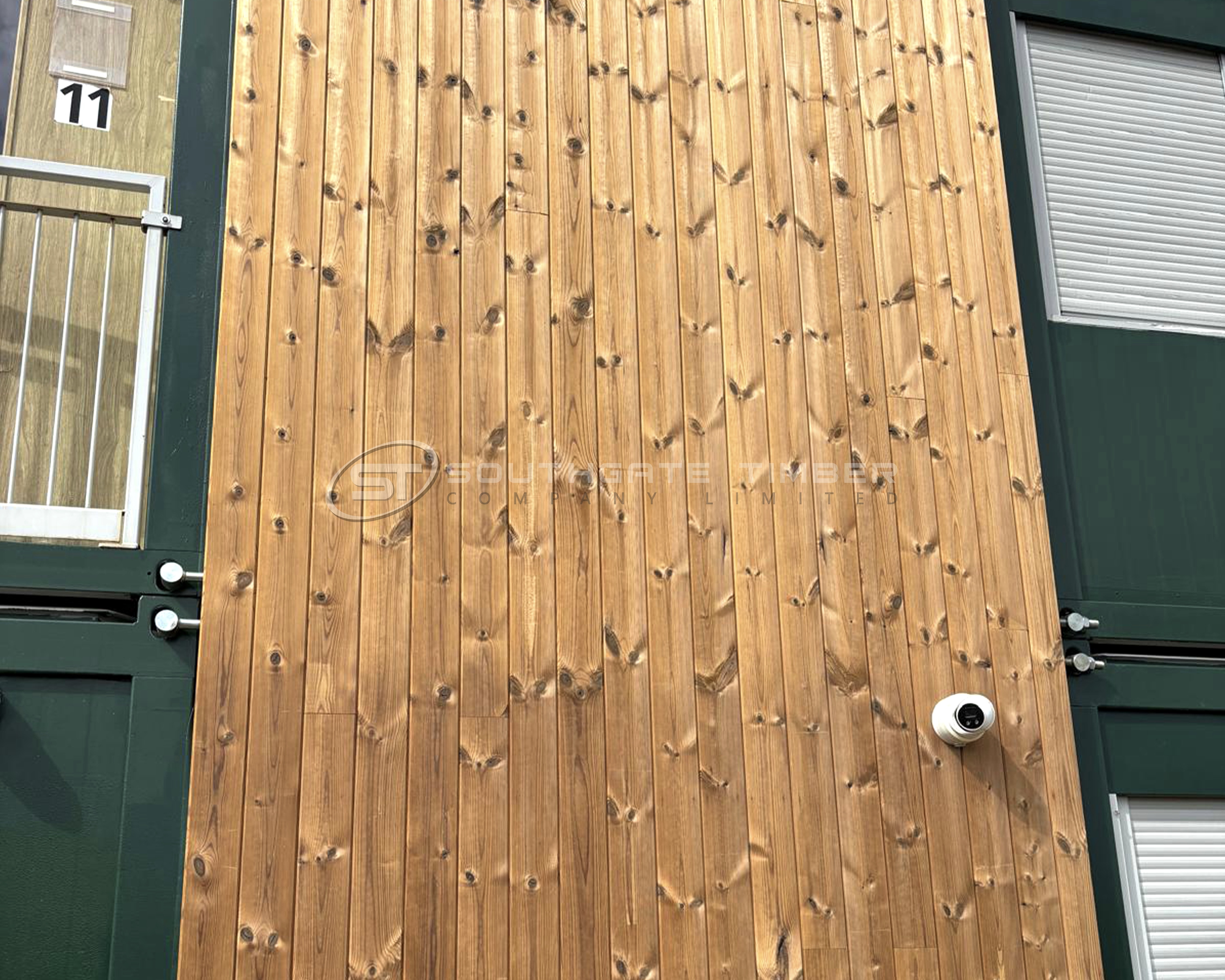 Exterior wall clad in Thermowood Pine 20 x 140mm Channel Groove Vertical ST6.TW, showing warm brown tones, visible knots, and clean vertical lines installed between dark green window frames. Thermally modified pine cladding with a modern vertical finish supplied by Southgate Timber.